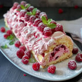 Une bûche roulée avec une crème rosée, décorée de feuilles de menthe et de framboises fraîches, posée sur un plat.